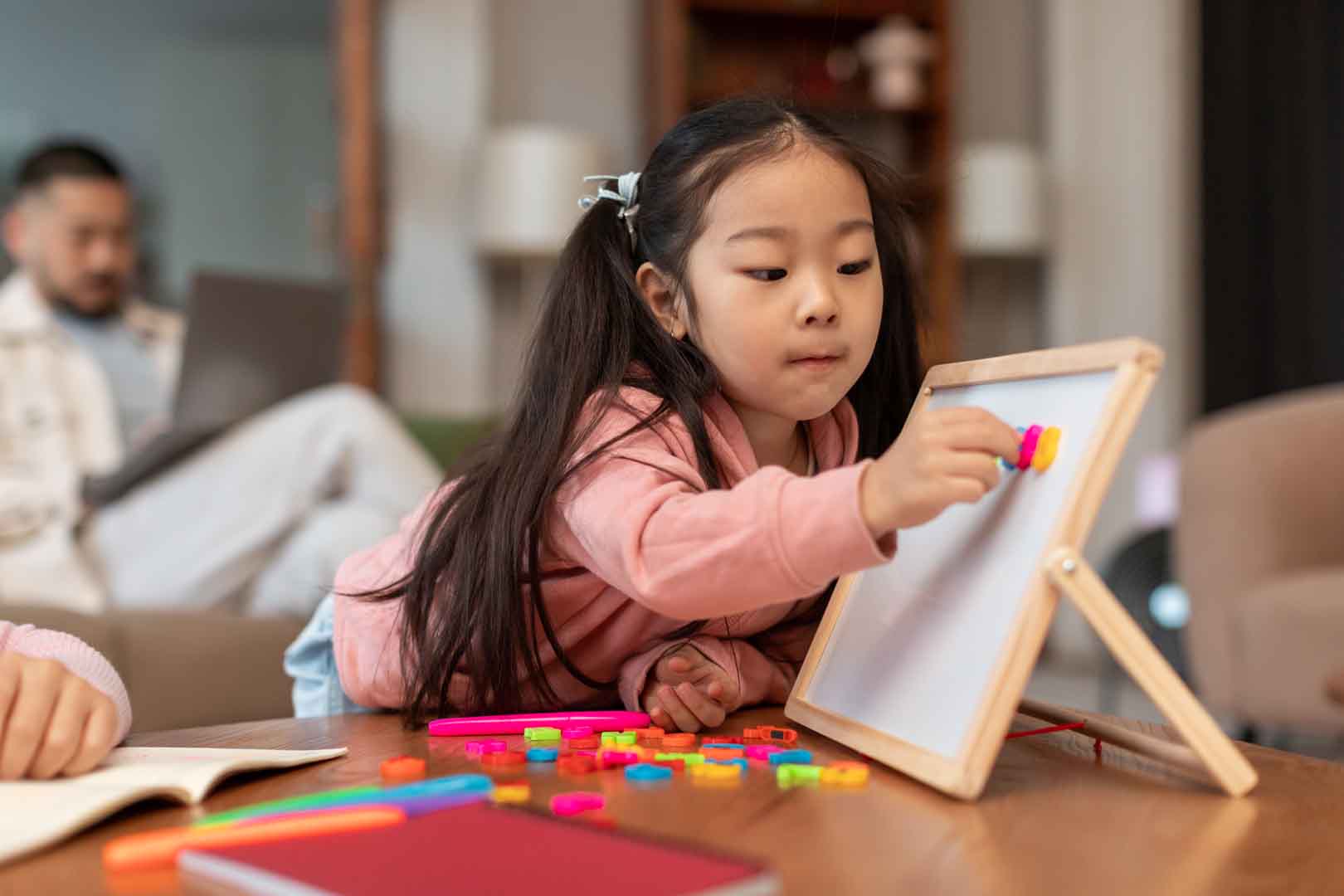 A young girl in a pink hoodie plays with colorful magnetic letters on a board, practicing early writing and literacy skills through fine motor activities.