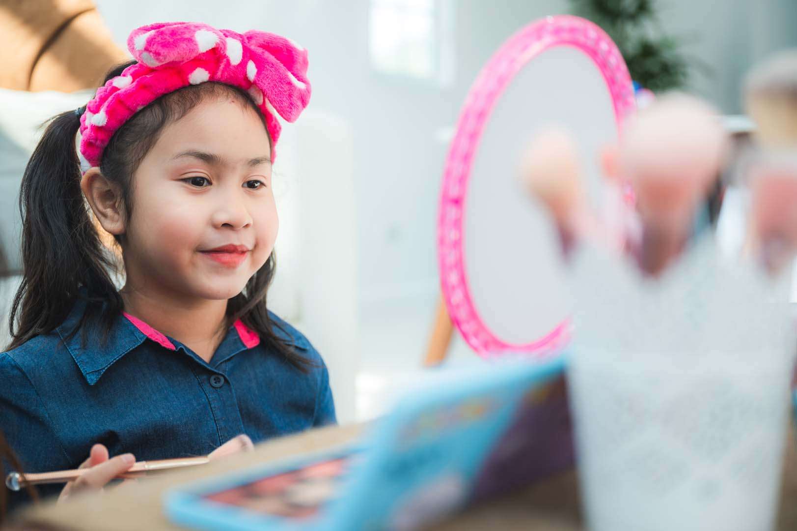 Young girl wearing a pink polka-dot headband practicing makeup in front of a mirror, engaging in imaginative play and self-expression.