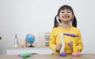 A smiling girl in a yellow shirt uses colorful play dough for a fun occupational therapy activity that strengthens hand muscles and creativity.