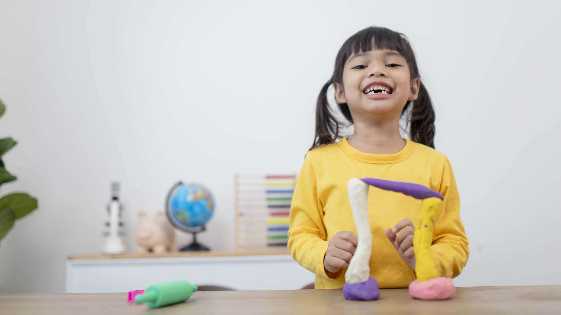 A smiling girl in a yellow shirt uses colorful play dough for a fun occupational therapy activity that strengthens hand muscles and creativity.