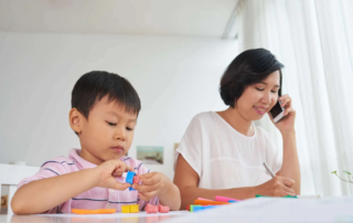 Young boy developing fine motor skills using therapy tools while mother supports from nearby during occupational therapy activity in Phnom Penh