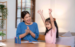 A young girl raises her arms in joy during a speech therapy session at OrbRom Center in Phnom Penh as her therapist claps in celebration.