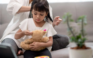 A young girl focuses on placing beads on a teddy bear’s ear while an adult helps style her hair in the background—supporting fine motor skills and focus.