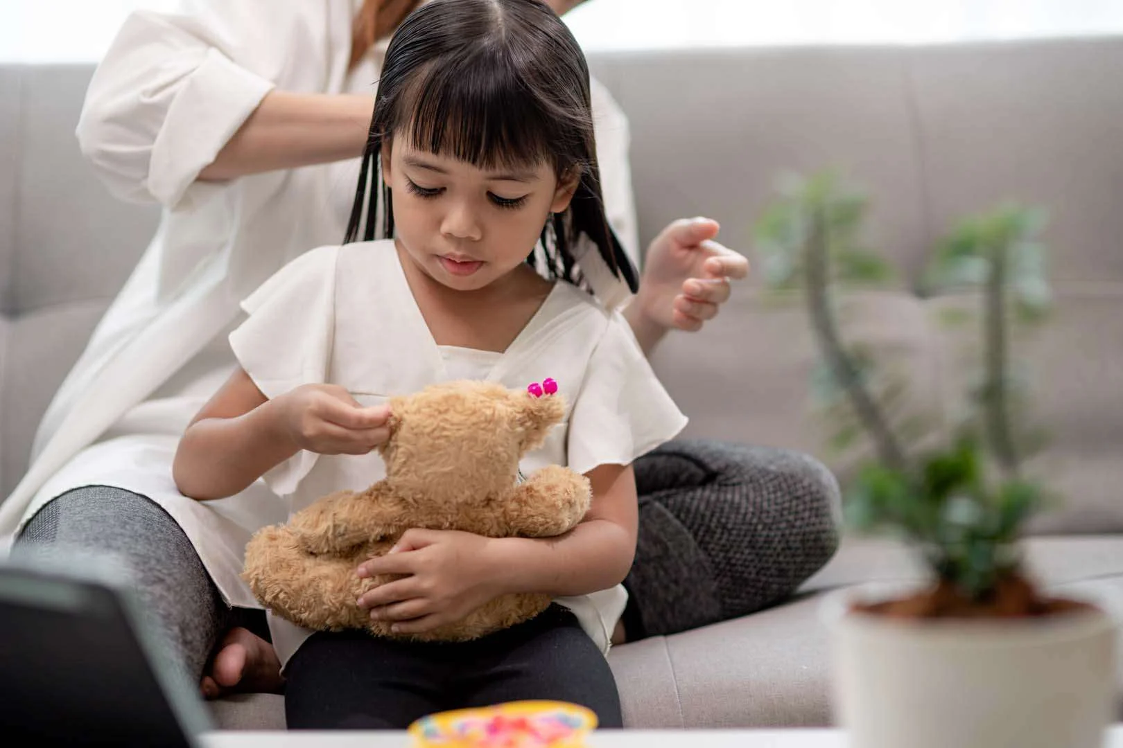 A young girl focuses on placing beads on a teddy bear’s ear while an adult helps style her hair in the background—supporting fine motor skills and focus.