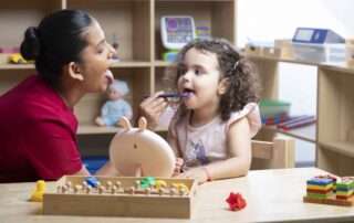 A speech therapist helps a young child strengthen tongue movement using a mirror and tongue depressor tool during therapy.