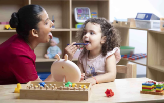 A speech therapist helps a young child strengthen tongue movement using a mirror and tongue depressor tool during therapy.