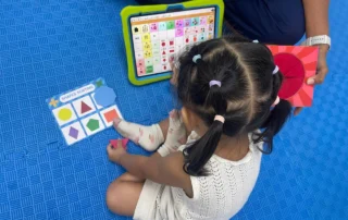 Child using AAC tablet during bilingual speech therapy session with mystery box learning activity
