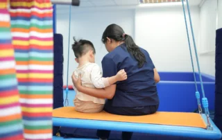 Child and occupational therapist sitting together on therapy swing for sensory regulation and body awareness.